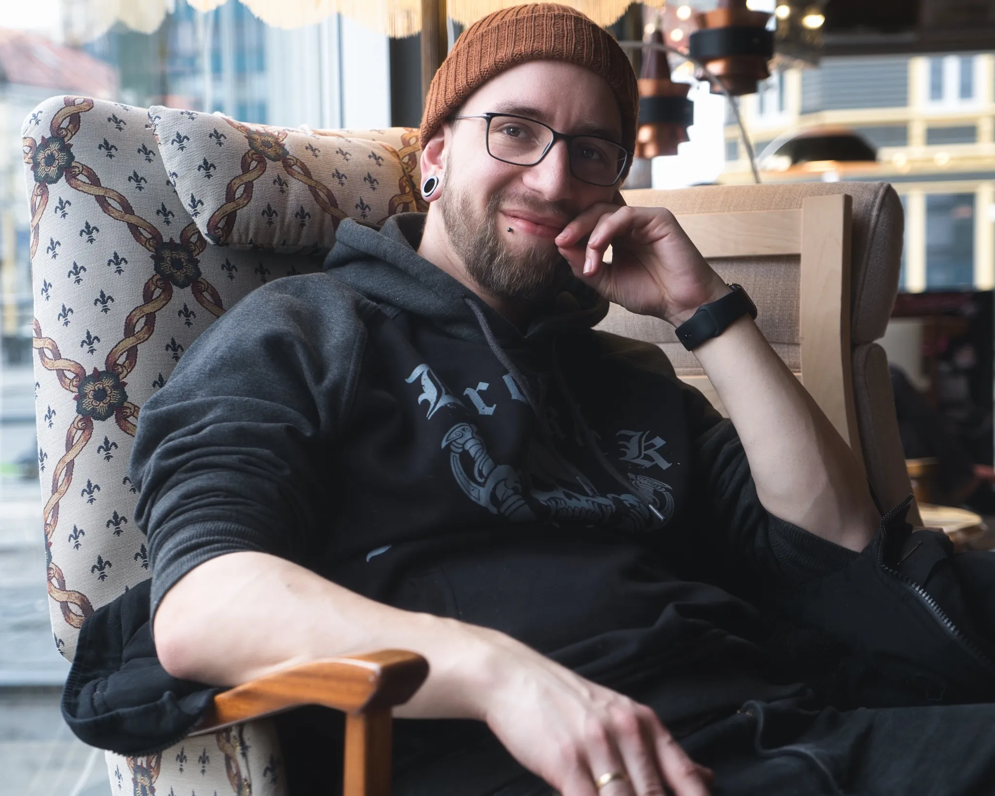 A young man with glasses and an orange beanie sits in a patterned armchair, wearing a dark hoodie. He has a beard and is smiling at the camera with his hand near his face.